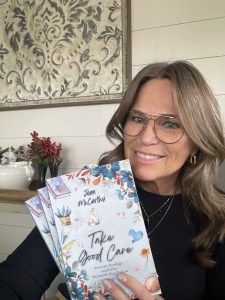 Author Jean McCarthy wearing glasses and a black shirt holding three copies of the book "Take Good Care"