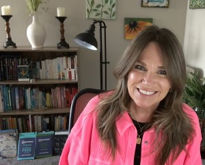 photo of UnPickled author Jean McCarthy wearing a pink shirt, seated next to a bookcase displaying a selection of her books and awards